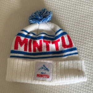 Vit och blå Minttu mössa med tofs - Snygg stickad mössa från Minttu i vitt med blå och röda detaljer. Mössan har blå ränder, stor röd text och en blåvit tofs på toppen. Framtill sitter en patch med Minttu-logga och bergmotiv. 