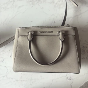 Beige handväska från Michael Kors - Snygg beige handväska från Michael Kors i stilren boxig modell. Väskan har både handtag och justerbar axelrem, samt dragkedja upptill. Tillverkad i strukturerat skinn med diskret logga framtill. Perfekt storlek för det viktigaste och passar till många outfits.