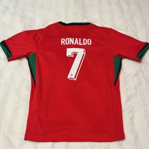 Portugal Ronaldo 7 fotbollströja röd) fotbollströja  - ✅ Fakta om tröjan: – Storlek: M (Medium) – Färg: Röd med gröna och svarta detaljer – Tryck: Ronaldo 7 på ryggen – Märke: Portugal landslagströja (inofficiell supportertröja) – Material: Lättviktig och ventilerande polyesterblandning – Skick: Begagnad men mycket välvårdad – inga hål, fläckar eller slitna sömmar – Tvättråd: Tvättas i låg temperatur (30°C), vänd ut och in för att skydda tryck  Den här tröjan är ett utmärkt köp för dig som följer Ronaldo, Portugal, eller bara älskar fotboll ⚽️ 