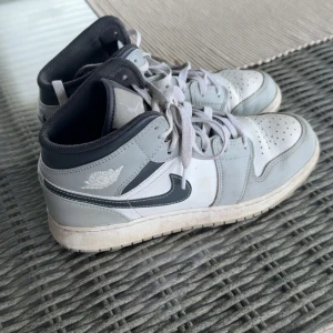 Nike Air Jordan 1 Mid grå/vit - Nike Air Jordan 1 Mid sneakers i grått och vitt med svarta detaljer och klassisk Jordan-logga på sidan. Skorna har snörning, perforerad tå och en bekväm platt sula.