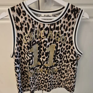 Leopardmönstrad basketlinne Chicago - Säljer en leopardmönstrad basketinspirerad linnetopp från No Logo med texten 'CHICAGO 11 DREAM TEAM' i guld och svart. Toppen är ärmlös med vita och svarta ribbade kanter och har en loose passform. Perfekt för dig som vill sticka ut med sportig vibe.
