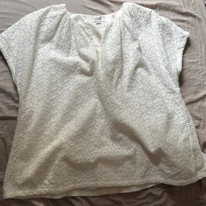 Vit broderad blus från COS - Superfin vit blus från COS i storlek 36. Blusen har korta ärmar, v-ringning med knappar och är täckt av broderade hålmönster som ger en luftig och somrig känsla. Perfekt till jeans eller kjol för en fräsch look. Stor i storlek. 
