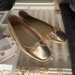 Guldiga ballerinaskor med rosett - Snygga ballerinaskor i metallic guld med fyrkantig tå och söt rosettdetalj framtill. Skorna har en platt sula och är tillverkade i syntetmaterial som ger en glansig finish. Aldrig använda 