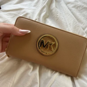 Beige plånbok från Michael Kors - Snygg beige plånbok från Michael Kors i skinn med stort guldfärgat MK-emblem på framsidan. Plånboken har dragkedja runtom, flera kortfack och ett myntfack med dragkedja. 