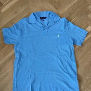 Ljusblå pikétröja från Ralph Lauren - Snygg ljusblå pikétröja från Polo Ralph Lauren i klassisk modell med kort ärm och broderad logga på bröstet. Tröjan har krage och knäppning framtill, tillverkad i bomull för en skön och luftig känsla. Perfekt till jeans eller shorts.