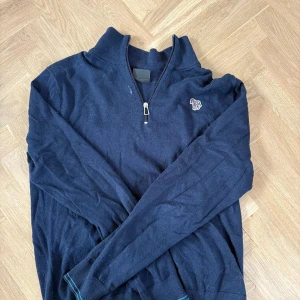 Mörkblå stickad tröja PAUL SMITH - Mörkblå stickad tröja med half zip och lång ärm. På bröstet finns en broderad zebra i färgglada ränder som cool detalj. Tröjan har ribbade muddar och hög krage, perfekt för kyligare dagar.