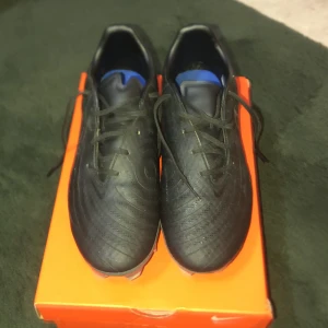 Nike phantom GX 2 Ac Black/Black 8,5 Nike det kommer med boxen och kvittot  - Säljer ett par svarta Nike fotbollsskor med vita detaljer och klassisk Swoosh på sidan. Skorna har snörning och slitstark syntetöverdel, samt dobbar för bra grepp på planen. Blå innersula för extra komfort. Perfekta för dig som vill ta spelet till nästa nivå.