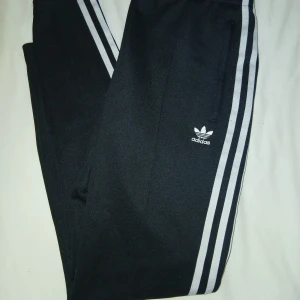 Svarta adidas originals träningsbyxor.  - Svarta Adidas originals byxor med klassiska vita ränder längs sidorna och broderad logga framtill. Byxorna har dragkedjeförsedda fickor och är gjorda i ett mjukt syntetmaterial. Perfekta för en sportig och avslappnad stil.