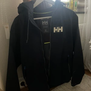 Mörkblå vindjacka från Helly Hansen - Mörkblå vindjacka  från Helly Hansen med stor HH-logga på bröstet. Jackan har huva, dragkedja framtill och praktiska fickor med dragkedja. Perfekt för blåsiga dagar och enkel att matcha med det mesta. Regular fit och sportig look.