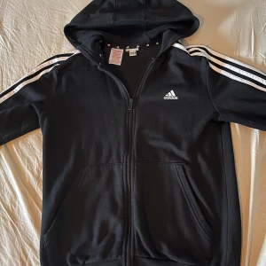 Svart Adidas tröja med dragkedja - Svart hoodie från Adidas med klassiska vita ränder längs ärmarna och Adidas-logga på bröstet. Tröjan har huva, dragkedja framtill och två fickor. Perfekt för en sportig och avslappnad stil. Nypris 499kr! 