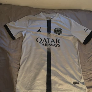 PSG Messi 30 Jordan fotbollströja - Vit PSG fotbollströja med svarta detaljer, Jordan-logga och Paris Saint-Germain-märke på bröstet. Svart vertikal rand framtill, Qatar Airways-tryck och Messi 30 på ryggen. Tillverkad i lätt och ventilerande material, perfekt för match eller träning.