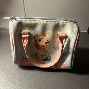 Frozen handväska med Elsa-motiv - Söt handväska med motiv av Elsa från Frozen, dekorerad med snöflingor, fjärilar och blommor. Väskan är ljusblå med rosa detaljer och handtag, har dragkedja upptill och är tillverkad i glansig syntet. 📩 Skicka gärna ett meddelande vid frågor eller om du vill ha flera bilder! 🌸😊 📦 Jag skickar snabbt och packar alltid med omsorg!