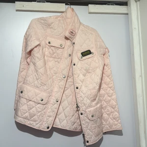 Ljusrosa quiltad jacka från Barbour - Quiltad jacka från Barbour i ljusrosa med snygga detaljer som silverfärgade knappar, dragkedja och flera fickor. Jackan har hög krage med spänne och Barbour-logga på bröstet. Perfekt för dig som vill ha en stilren och trendig look.Men bältet är borta 