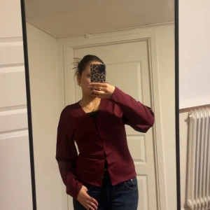 Vinröd kofta med knappar - Säljer en vinröd kofta med v-ringning och knäppning framtill. Modellen är figurnära med långa ärmar och enkel design, perfekt att slänga över ett linne eller t-shirt. Färgen är djup vinröd och materialet känns mjukt och bekvämt.