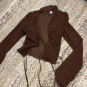 Brun blus/jacka från H&M Divided - Säljer en brun, transparent overshirt från H&M Divided i omlottmodell med långa ärmar och tunna knytband framtill. Perfekt att slänga över ett linne. Storlek XS