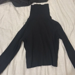 Svart ribbad polotröja med pärlor - Svart ribbad polotröja med lång ärm och höga muddar. Ärmarna är dekorerade med snygga pärlor längs kanten för en extra touch. Tröjan är tight i modellen och har en hög polokrage som ger en clean och stilren look.