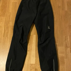 Svarta vindbyxor från Haglöfs - Svarta vindbyxor från Haglöfs med elastisk midja och reflexdetaljer vid bensluten. Perfekta för kalla dagar och utomhusaktiviteter. Lätta att dra på och av, med diskret Haglöfs-logga på benet.