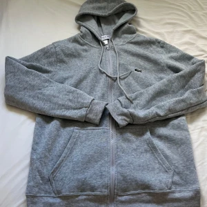 Grå hoodie från Lacoste med dragkedja - Snygg grå hoodie från Lacoste med klassisk krokodillogga på bröstet. Tröjan har huva med snörning, två stora fickor fram och hel dragkedja. Regular fit och tillverkad i mjuk bomullsmix, perfekt för chill dagar.