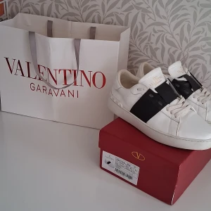 Valentino Garavani vita sneakers - Snygga vita sneakers från Valentino Garavani med bred svart rem över vristen och klassisk logga på plösen. Skorna har snörning, platt sula och coola nitar baktill. Tillverkade i skinn för en lyxig känsla och stilren look. Allting medföljer förutom kvitto