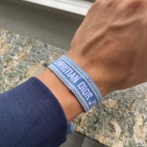 Blått Christian Dior J'Adior armband - Snyggt armband från Christian Dior i ljusblått tyg med broderad text 'CHRISTIAN DIOR J'ADIOR' i vitt. Armbandet har justerbar knytning med två tofsar i ändarna för en trendig look. Perfekt accessoar för att lyfta din outfit.