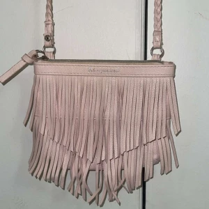 Ljusrosa axelväska med fransar - Supersöt ljusrosa axelväska i skinnimitation med långa fransar framtill och flätat axelband. Väskan har dragkedja upptill och en stilren, mjuk form. Perfekt för dig som vill ha en boho-inspirerad look och gillar detaljer som sticker ut.
