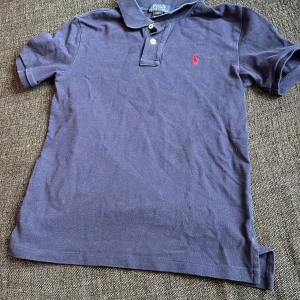 Marinblå pikétröja från Polo Ralph Lauren - Klassisk marinblå pikétröja från Polo Ralph Lauren med korta ärmar och krage. Tröjan har två knappar vid halsen och en röd broderad logga på bröstet. Perfekt för en stilren och avslappnad look.