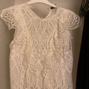 Vit spetsblus med volangdetaljer - Superfin vit blus i spets med vackra broderade mönster och volangdetaljer på axlarna. Ärmlös modell med rund halsringning och genombruten design som ger en romantisk och trendig look. Perfekt för dig som vill sticka ut med en unik topp.