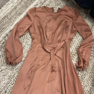 Rosa långklänning från SHEIN - Supersnygg rostbrun/rosa satin långklänning från SHEIN med långa ärmar, knytband i midjan och lätt omlott framtill. Stängs med dragkedja i ryggen. Storlek S