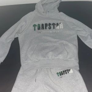 Trapstar dress - Säljer en grå hoodie från Trapstar med coolt tryck i grönt, svart och vitt på bröstet. Tröjan har huva med dragsko och en stor magficka. Perfekt för dig som gillar streetwear och vill ha en avslappnad stil.