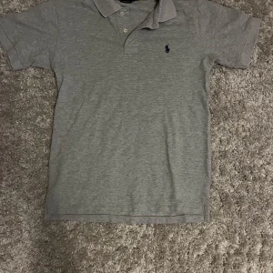 Grå pikétröja från Polo Ralph Lauren - Snygg grå pikétröja från Polo Ralph Lauren i bomull. Klassisk krage, korta ärmar och två knappar framtill. Broderad marinblå logga på bröstet. Modellen är slim fit och passar perfekt till en clean och stilren look.