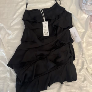 Satin klänning med volang - Supersnygg svart satin kortklänning med flera lager volanger och smala axelband. Med prislapp kvar vid snabb affär kan priset sänkas💕