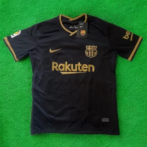 FC Barcelona svart Nike matchtröja - Snygg FC Barcelona matchtröja från Nike i svart med guldiga detaljer och tryck. Tröjan har korta ärmar, V-ringad krage och är tillverkad i lätt, ventilerande Dri-FIT-material. Officiella loggor från både klubben och sponsorerna Rakuten och UNICEF.