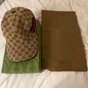 Gucci keps med GG-mönster och ränder - Exklusiv keps från Gucci i beige och brunt med klassiskt GG-monogram över hela kepsen. Sidan pryds av gröna och röda ränder. Kepsen är tillverkad i bomullscanvas och har justerbar rem baktill. Perfekt för dig som vill sticka ut med lyxig streetstyle.