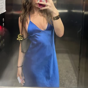 Blå satinklänning med smala axelband och öppet rygg - Säljer en snygg blå satinklänning med smala axelband och djup v-ringning. Klänningen har en slät, glansig yta och faller fint längs kroppen. Perfekt för dig som gillar en stilren och trendig look.
