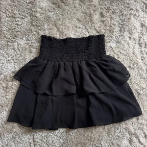Svart volangkjol - Supersöt svart volangkjol från Chelsea med smockad midja och dubbla volanger. Säljer då jag föredrar att använda shorts, och tycker det känns tråkigt att låta en kjol i detta skick bara ligga. Jag hoppas att denna kan få ett nytt hem, någonstans där den inte enbart får ligga i garderoben🥹🥰