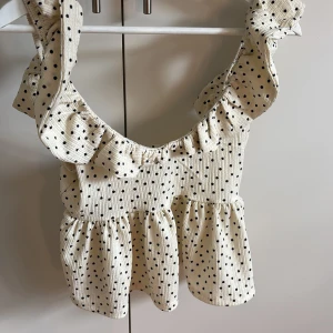 Prickig volangtopp med breda axelband - Supersöt topp i ljusbeige med svarta prickar och breda volangprydda axelband. Toppen har en smockad, ribbad struktur och volang nertill för extra flowy känsla. Perfekt för dig som gillar romantisk och lekfull stil.