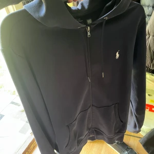 Marin blå hoodie från Polo Ralph Lauren - Marin blå hoodie från Polo Ralph Lauren med dragkedja framtill, huva med snörning och två stora fickor. Klassisk vit logga broderad på bröstet. Tillverkad i mjuk bomull, perfekt för chill dagar eller när du vill ha en clean look. Ny pris 1800!!