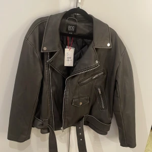 Svart oversized skinnjacka från Urban Outfitters - Säljer en svart skinnjacka från BDG Urban Outfitters i . Jackan är oversized med bälte i midjan och quiltat innerfoder. Perfekt för dig som vill ha en edgy och klassisk look. Storlek XS men eftersom den är oversized passar den även större storlekar upp till M/L. Helt oanvänd med prislapp kvar och nypris var 999kr. Hör av dig vid funderingar!❣️