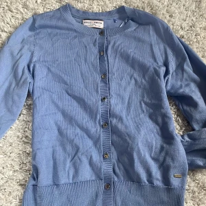 Ljuslila/blå  kofta från Holly & Whyte - Supersöt ljusblå kofta från Holly & Whyte med  snygga guldknappar  knappar framtill. Långärmad och ribbade muddar vid ärmslut och nederkant. Perfekt för att göra en basic outfit roligare!