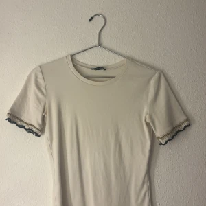Beige topp med spets och guldtråd - Snygg beige topp med korta ärmar och rund halsringning. Ärmsluten har dekorativa detaljer med spets, guldtråd och svarta kanter som ger en unik och trendig look. Perfekt för dig som gillar subtila men coola detaljer.