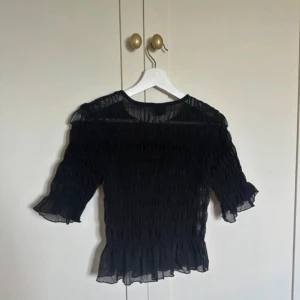 Svart volangblus från Gina Tricot - Supersöt svart blus från Gina Tricot i transparent, lätt material. Blusen har smockad struktur, volangkant nertill och vid ärmslut samt rund halsringning. Perfekt för dig som gillar trendiga detaljer och en feminin look.