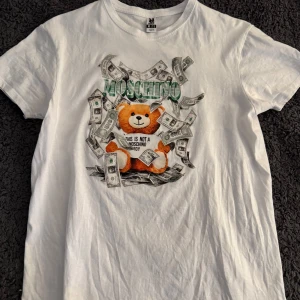 Vit t-shirt med nalle och dollartryck - Vit t-shirt från Roly i bomull med coolt tryck framtill: en nallebjörn omgiven av dollarsedlar och texten 'MOSCHINO'. Klassisk passform och rund hals, perfekt för dig som gillar statement-plagg.