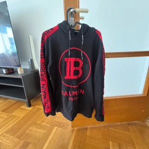 Svart och röd Balmain hoodie - Svart hoodie från Balmain med röda detaljer och stor logga på bröstet och på ärmarna. Tröjan har huva med snörning, långa ärmar och dragkedjor vid ärmsluten. Storlek L men passar M (HELT NYTT SKICK)