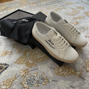 Saint Laurent vita sneakers i canvas - Snygga vita sneakers från Saint Laurent i canvas med beige sula och handskriven logga på sidan. Klassisk låg modell med rund tå, snörning och diskreta detaljer. Perfekta för dig som gillar stilren och lyxig streetwear.helt nya och oanvända 