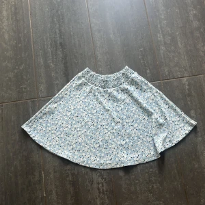 Blommig blå kortkjol från 157 XS - Supersöt ljusblå kortkjol från 157 med vita och svarta småblommor. Kjolen har elastisk midja för extra bekvämlighet och är perfekt för varma dagar. 