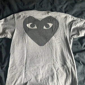 Grå Comme des Garçons Play t-shirt - Säljer en grå t-shirt från Comme des Garçons Play med stort svart hjärta med ögon på ryggen och klassisk PLAY-logga framtill. Storleke är L men sitter mer som M