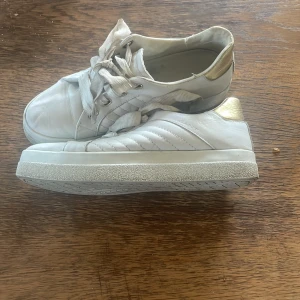Vita sneakers från Gant med gulddetalj - Snygga vita sneakers från Gant med breda snören och guldfärgade detaljer på hälen. Skorna har en klassisk rund tå, platt sula och är tillverkade i skinn. Perfekta för dig som gillar stilrena och fräscha sneakers med en touch av guld. se sista bilden på defekterna annars fint skick💕