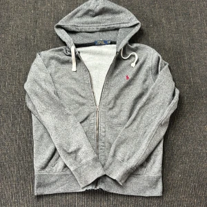 Grå hoodie från Polo Ralph Lauren  - Snygg grå hoodie från Polo Ralph Lauren med dragkedja och vit snörning i huvan. Klassisk röd logga broderad på bröstet. Mjuk insida och praktiska fickor framtill. Perfekt för chill dagar eller när du vill ha en clean streetstyle. Skicket är toppen! Hör gärna av er vid frågor eller funderingar