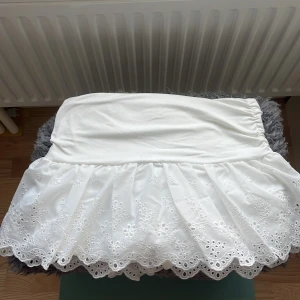Vit kortkjol med broderade spetsdetaljer - Supersöt vit kortkjol med broderade blommor och spetskant. Kjolen har elastisk midja och inbyggda shorts under för extra komfort. Perfekt för sommarens varma dagar och ger en romantisk vibe till din outfit.