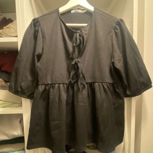 Svart blus med puffärm och knyt - Svart blus från lager 157 med puffiga korta ärmar och två knytningar framtill. Modellen är lös och har en volang nedtill som ger en flowy känsla. Perfekt för dig som gillar en söt och trendig look med extra volym i ärmarna.